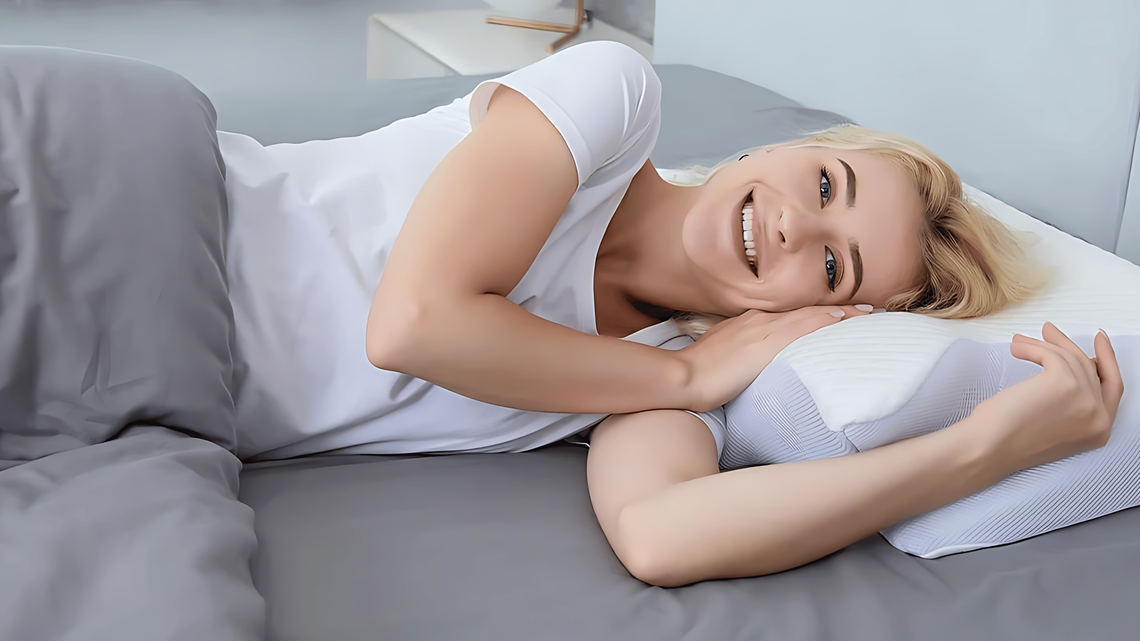 Smiling blonde woman lying on her side in bed, resting her head on a contoured pillow. She's wearing a white t-shirt and appears comfortable and relaxed. The bedding is gray and the pillow has a white design.